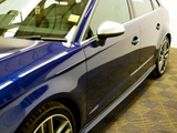 A shiny blue car is parked, showcasing a sleek design with a side mirror and a reflection on the side panel. It is indoors on a patterned floor. There is a yellow sticker with 