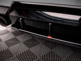 A black car's rear bumper features an aerodynamic diffuser with two vertical fins and a red logo. It rests on a patterned rubber matting surface in an indoor space.