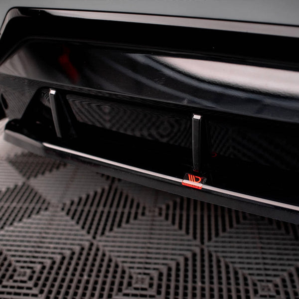 A black car's rear bumper features an aerodynamic diffuser with two vertical fins and a red logo. It rests on a patterned rubber matting surface in an indoor space.