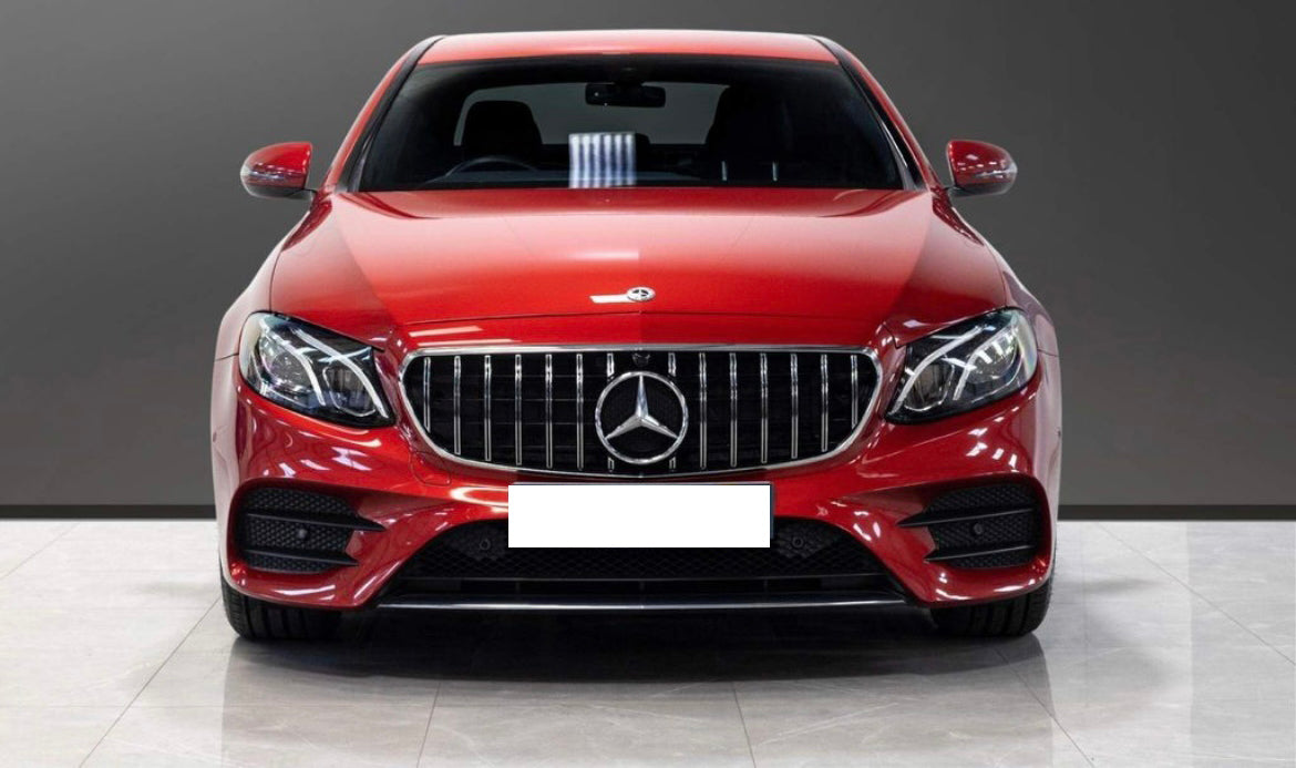A red sedan is parked indoors, featuring a prominent grille with a silver emblem at the center, surrounded by sleek headlights. The floor is tiled and the background is plain gray.