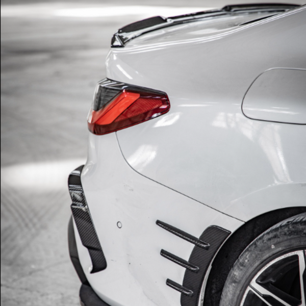 The image shows the rear corner of a white car, with a visible taillight and aerodynamic features. It's parked in a spacious, gray indoor environment.