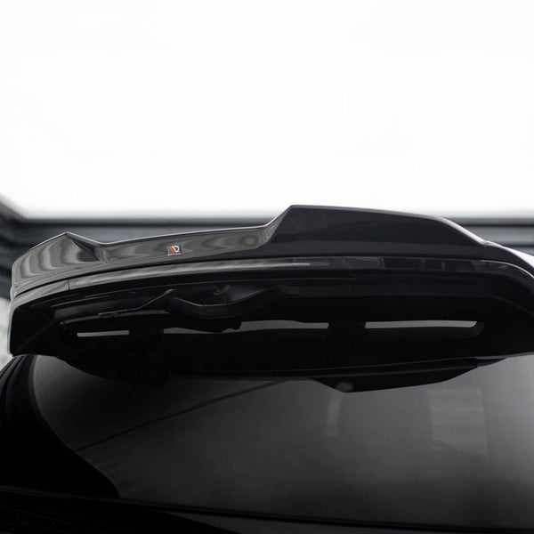 A shiny black car's rear spoiler is raised, highlighting its glossy finish. In a dimly lit garage setting, a small red and black "V" logo is visible on the spoiler.