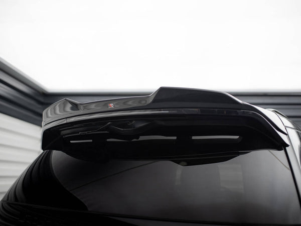 A shiny black car's rear spoiler is raised, highlighting its glossy finish. In a dimly lit garage setting, a small red and black "V" logo is visible on the spoiler.