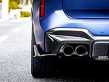 A blue car's rear with dual exhaust pipes and carbon fiber detailing is stationary on a paved road; blurred background suggests an urban environment.