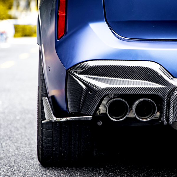 A blue car's rear with dual exhaust pipes and carbon fiber detailing is stationary on a paved road; blurred background suggests an urban environment.