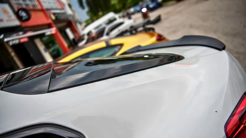 A white car's rear end with a spoiler is parked on a street, surrounded by other vehicles and buildings. The street has a relaxed urban environment.