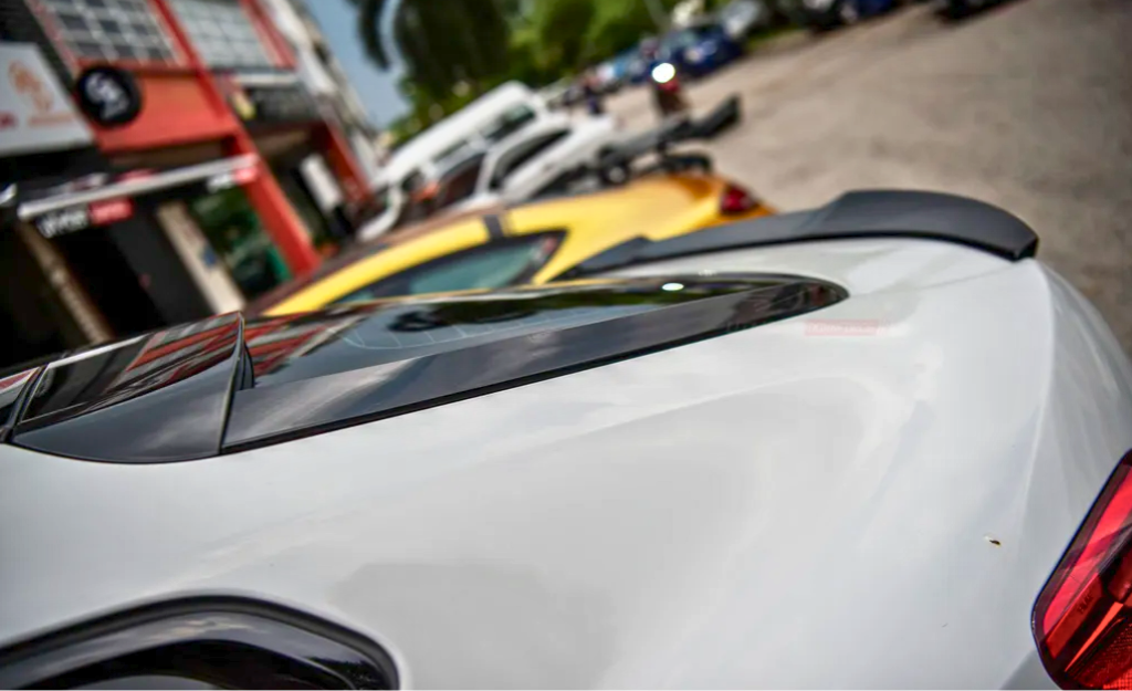 A white car's rear end with a spoiler is parked on a street, surrounded by other vehicles and buildings. The street has a relaxed urban environment.