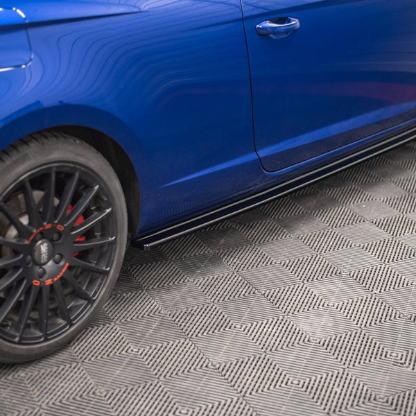 A blue car is parked on a textured gray floor. It features black wheels with red accents, and the side panel is visible with lines reflecting overhead lights.
