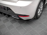 The rear of a silver car with a visible tire, exhaust pipe, and red reflector rests on a textured gray, grid-patterned surface. Partial signage reads 