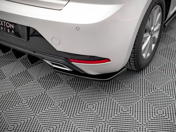 The rear of a silver car with a visible tire, exhaust pipe, and red reflector rests on a textured gray, grid-patterned surface. Partial signage reads "XTON Design."