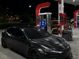 A sleek black car is parked under bright lights at a gas station, near fuel pumps and trash bins, with a nighttime backdrop of trees and distant buildings.