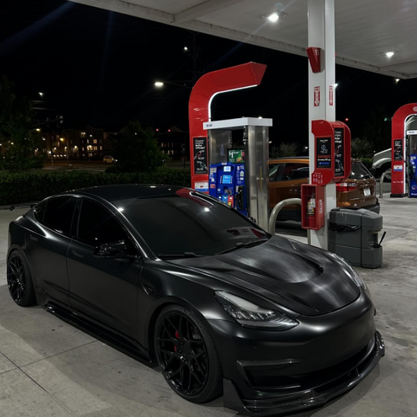 A sleek black car is parked under bright lights at a gas station, near fuel pumps and trash bins, with a nighttime backdrop of trees and distant buildings.