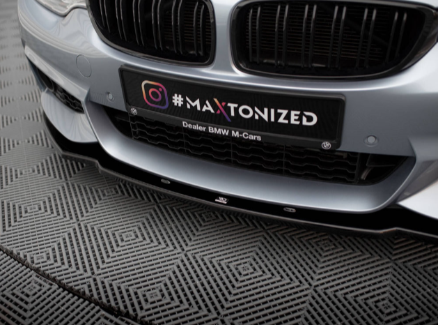 A car with a distinct grille is parked on a textured surface. The license plate reads "#MAXTONIZED Dealer BMW M-Cars," featuring an Instagram logo on the left.