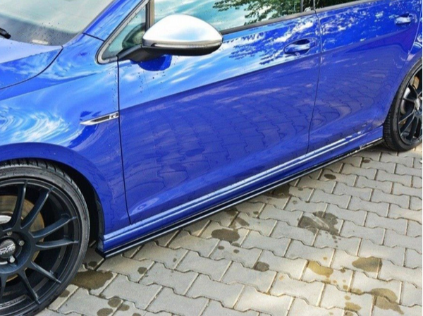 A blue car is parked on a wet, paved surface. The reflection of trees can be seen on the car's shiny surface, and black rims are visible on the wheels.
