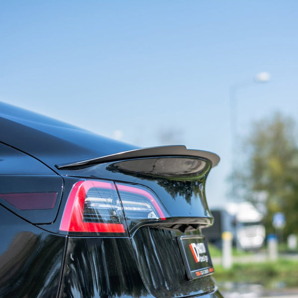 A sleek black car with a prominent rear spoiler is parked outdoors, with clear taillights visible. Trees and sky form the background, providing a serene setting.