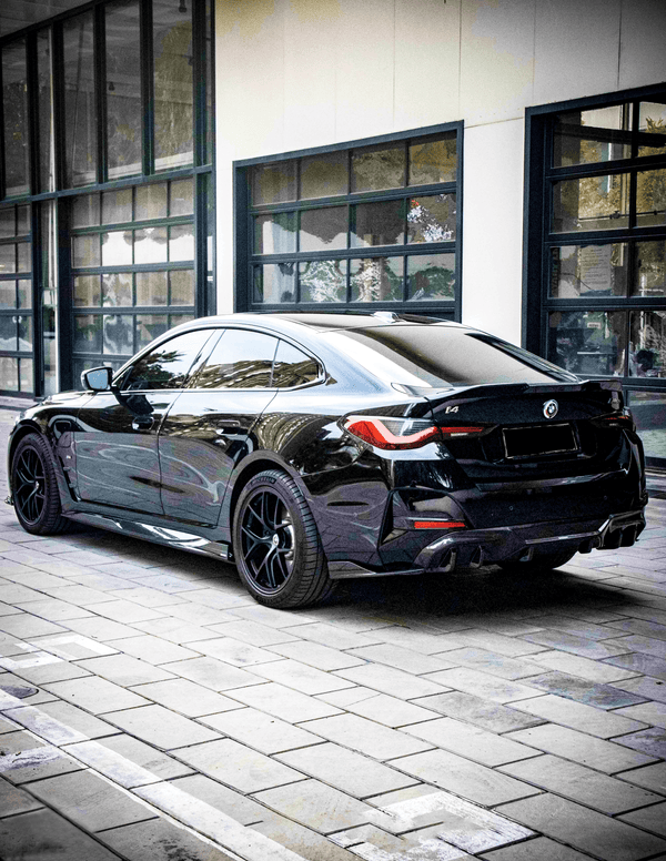 A sleek black BMW i4 is parked on a tiled urban street, reflecting nearby buildings in its shiny surface, against a backdrop of large windows.