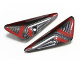 Two carbon fiber car parts with red accents and holes, positioned on a white surface. Each piece is triangular with a glossy finish, showing a textured pattern.