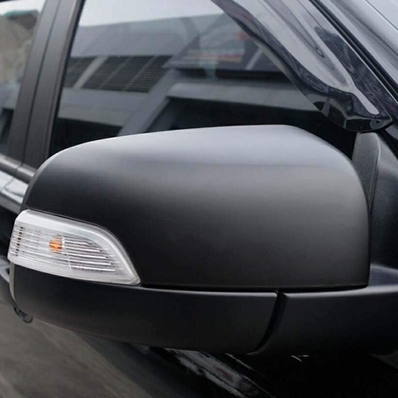 A black car side mirror with an integrated turn signal is attached to a vehicle's door, reflecting an urban setting.