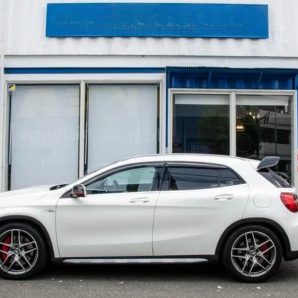 A white SUV is parked in front of a white and blue building with a partially visible sign above. Red brake calipers and alloy wheels accent the vehicle.