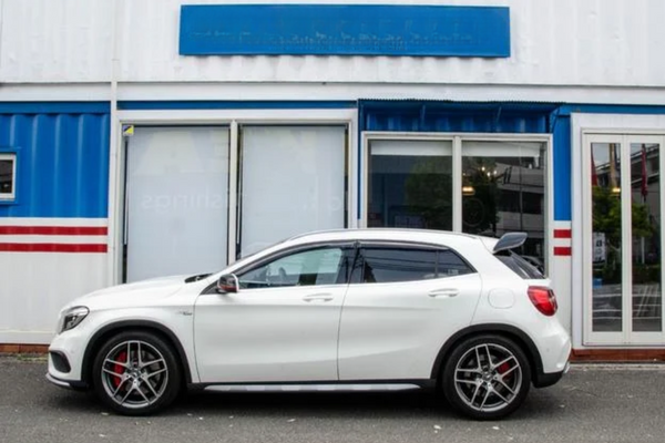 A white SUV is parked in front of a white and blue building with a partially visible sign above. Red brake calipers and alloy wheels accent the vehicle.