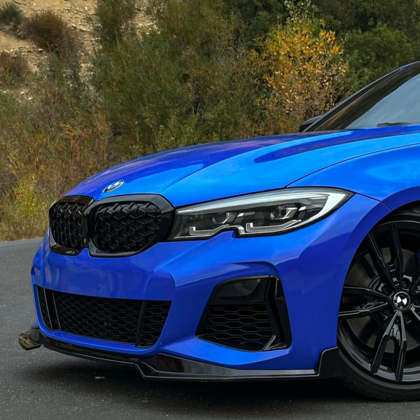 A blue car with a prominent grille and sleek headlights is parked on a road, surrounded by trees and shrubs. The shiny black wheel is partially visible.