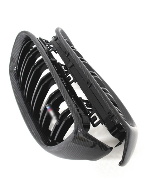 A shiny, carbon-fiber car grille featuring multiple vertical slats is angled upward on a plain white background, indicating its readiness for vehicle installation.