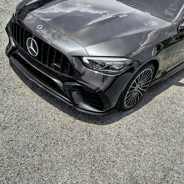 A sleek, dark-colored Mercedes car is parked on a gravel surface, highlighting its prominent grille and intricate wheel design under a partly cloudy sky.