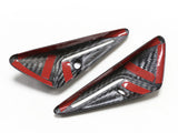 Two black carbon fiber trim pieces with red accents rest on a white surface; each has a small circular hole.