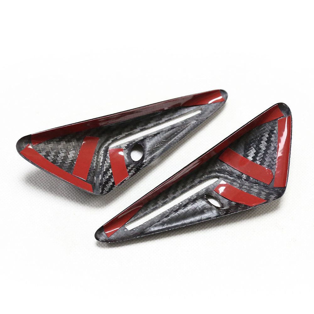 Two black carbon fiber trim pieces with red accents rest on a white surface; each has a small circular hole.