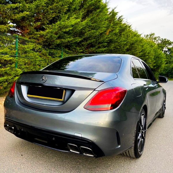 A silver Mercedes-Benz sedan is parked on the street, facing away. It's alongside a tall, green hedge under a cloudy sky, with no visible text.