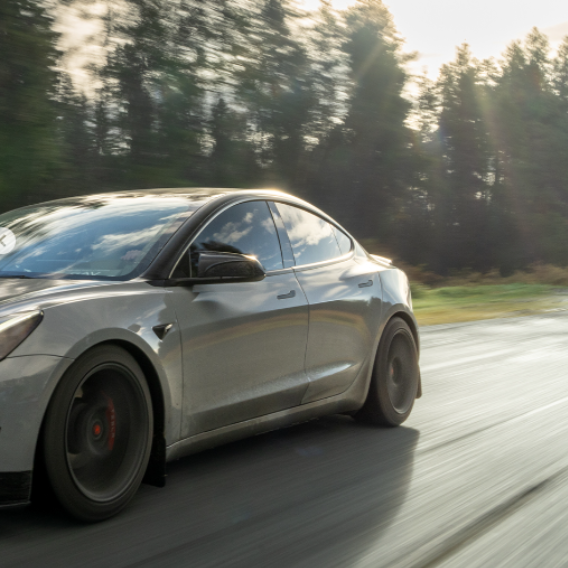 A sleek silver car speeds down a road, surrounded by a blurred forest, with sunlight peeking through the trees.