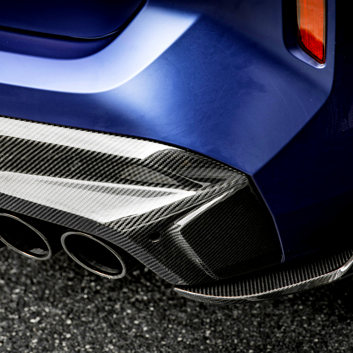 A car's blue rear bumper features a glossy carbon fiber diffuser with three exhaust pipes, parked on an asphalt surface. The setting suggests a modern automotive design.