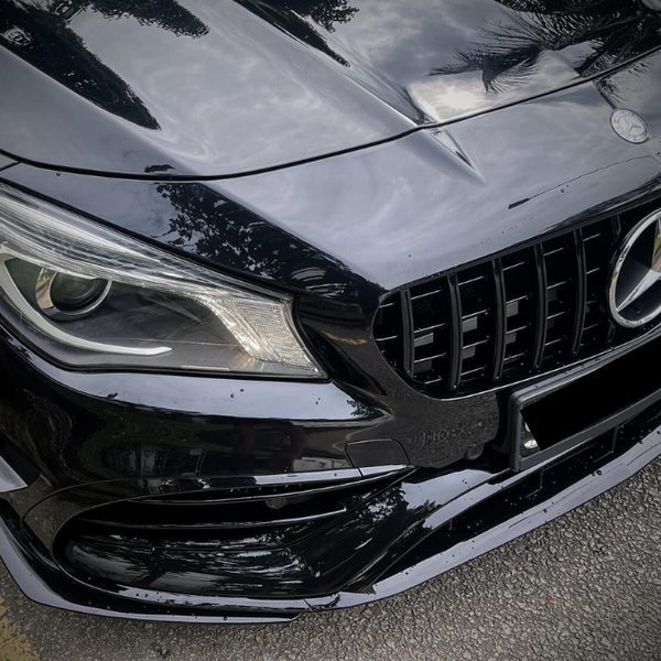 A black car's front end with a prominent grille and headlight is parked on a street, reflecting trees and clouds, featuring a three-pointed star emblem on the hood.