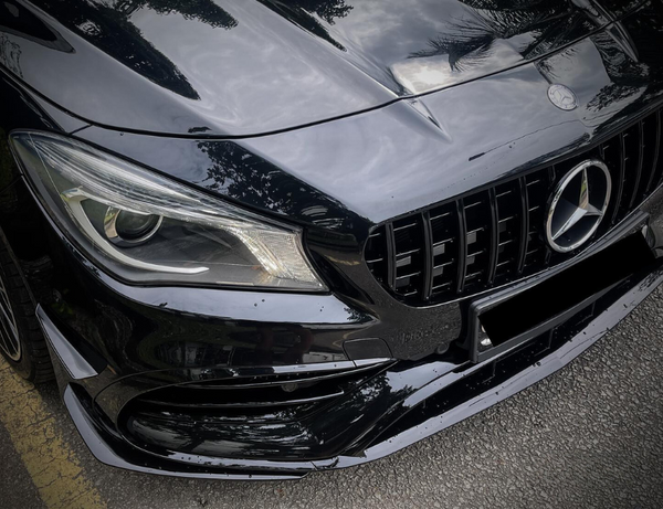 A black car's front end with a prominent grille and headlight is parked on a street, reflecting trees and clouds, featuring a three-pointed star emblem on the hood.