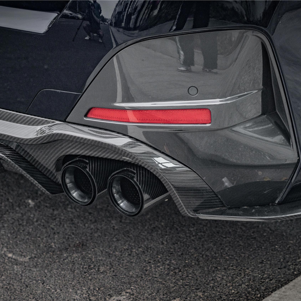 A car's rear bumper features dual exhaust pipes and a carbon fiber diffuser. It's parked on asphalt with a visible red reflector strip.