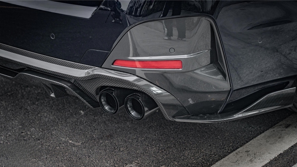A car's rear bumper features dual exhaust pipes and a carbon fiber diffuser. It's parked on asphalt with a visible red reflector strip.