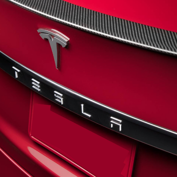 A red car features a prominent Tesla emblem and lettering on the rear, with a carbon-fiber trim above.