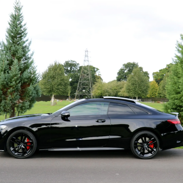 A sleek black car is parked on a paved road, surrounded by green grass and tall trees, under a partly cloudy sky.
