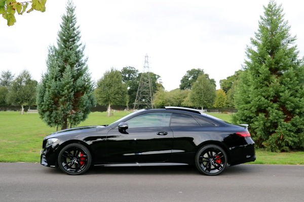 A sleek black car is parked on a paved road, surrounded by green grass and tall trees, under a partly cloudy sky.