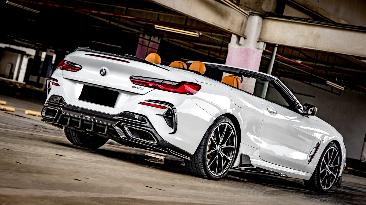 A white convertible sports car, displaying sleek design and chrome wheels, is parked in a dimly lit indoor structure. "BMW" badge and "840i" insignia visible on the rear.