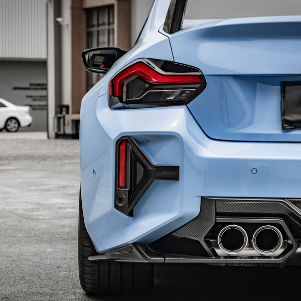 A blue car's rear is parked in a lot, featuring prominent exhausts and taillights, with a white car and industrial building in the blurred background.