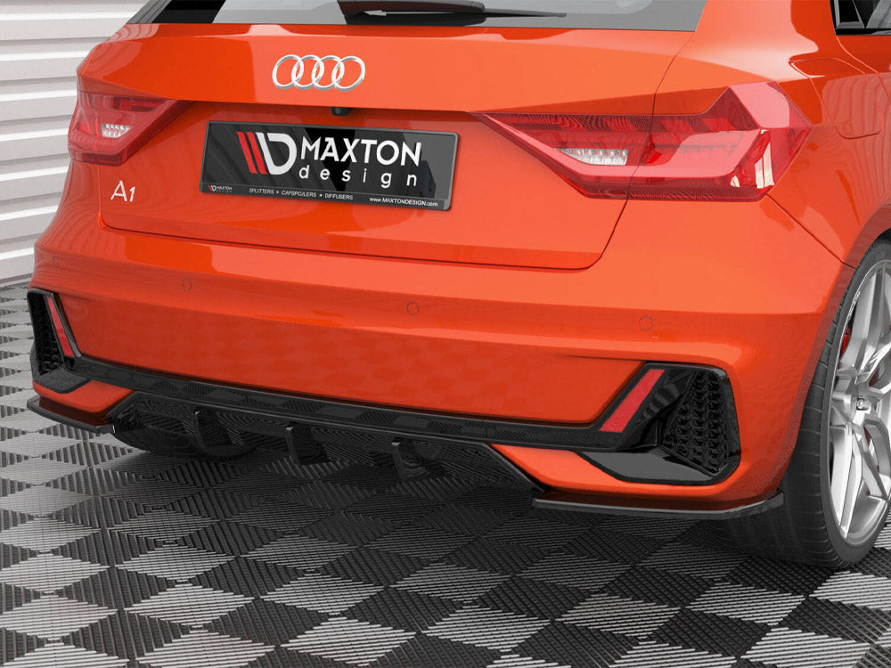 A bright orange Audi A1 is parked on a checkered floor, displaying a Maxton design rear spoiler and diffuser. The license plate reads "MAXTON design" with mentions of "SPLITTERS," "CAPSPOILERS," "DIFFUSERS," and "WWW.MAXTONDESIGN.COM."