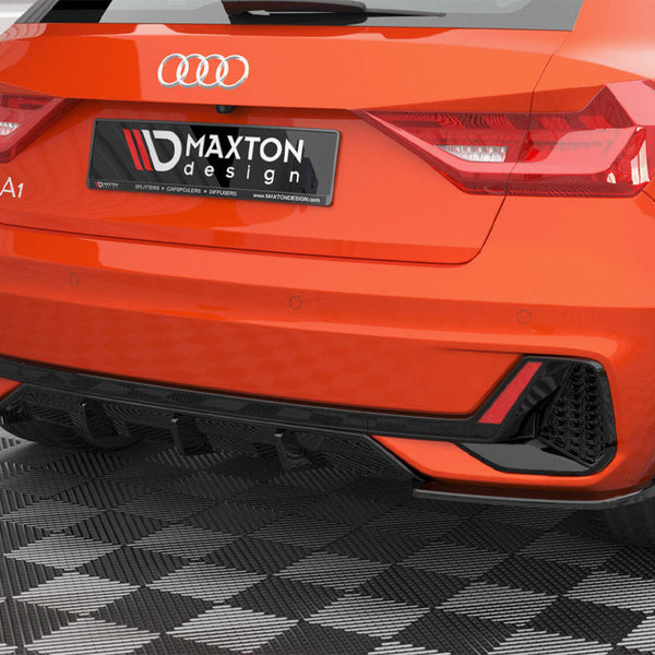 A bright orange Audi A1 is parked on a checkered floor, displaying a Maxton design rear spoiler and diffuser. The license plate reads "MAXTON design" with mentions of "SPLITTERS," "CAPSPOILERS," "DIFFUSERS," and "WWW.MAXTONDESIGN.COM."