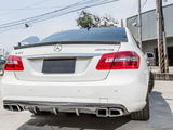 A white Mercedes-Benz E 63 AMG is parked on a concrete surface. It features quad exhausts and is located near a gray industrial building with overhead power lines.