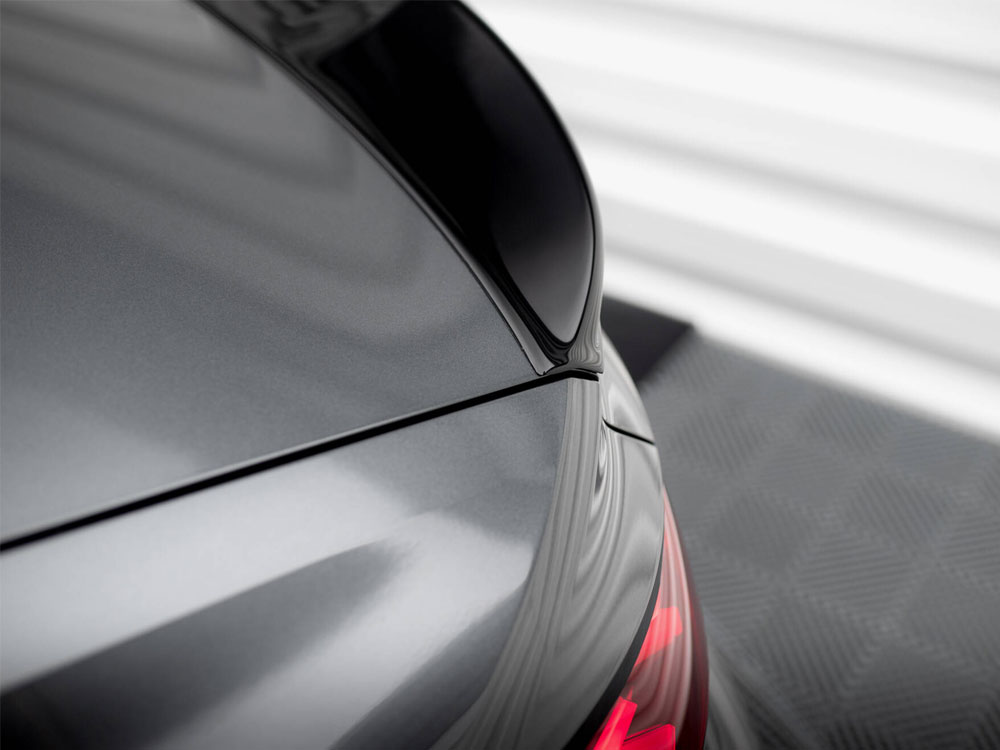 A shiny car's rear section with a sleek black spoiler, partially visible red taillights, is parked on a checkered pattern floor, set against a blurred, light-colored background.