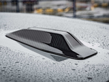A carbon fiber car roof scoop sits on a wet surface, covered in raindrops. The background is blurred, suggesting an outdoor setting.