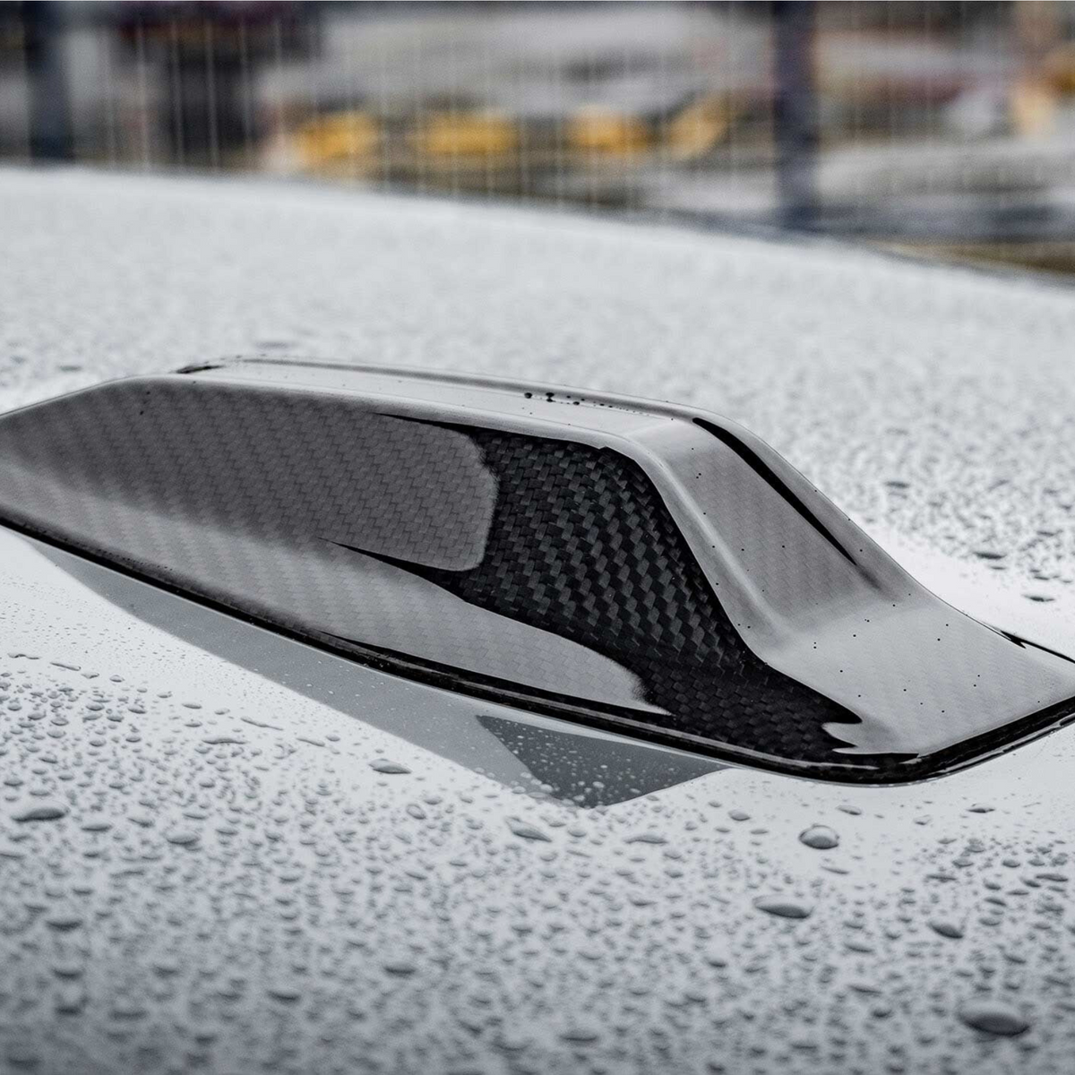 A carbon fiber car roof scoop sits on a wet surface, covered in raindrops. The background is blurred, suggesting an outdoor setting.