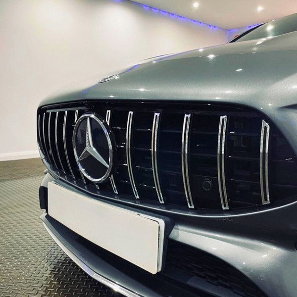 Car front with Mercedes-Benz logo on grille, parked in a lit indoor space with a metallic floor and white walls. A white license plate is attached below the grille.