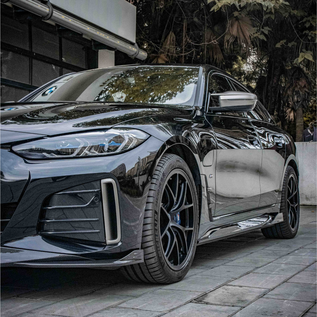 A sleek, black car is parked on a tiled pavement. The setting includes a modern building on the left and lush trees in the background, creating an urban yet natural ambiance.