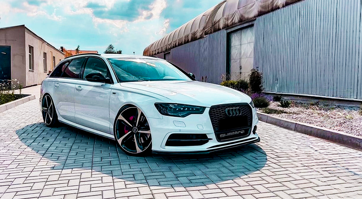 A white Audi car is parked on a paved driveway. The car is stationary, with a building and greenery in the background. The license plate reads "QUATTRO."